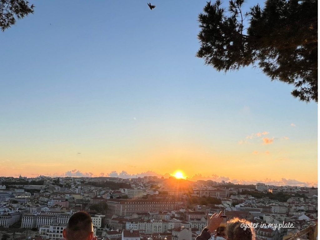A wide shot of the Lisbon cityscape at sunset, with the sun low on the horizon, casting an orange glow over the red-tiled roofs and distant buildings, seen from a high viewpoint with silhouetted trees and people in the foreground