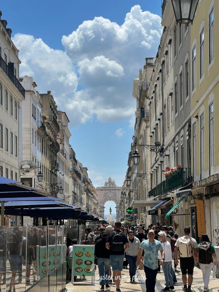 A bustling pedestrian street (Rua Augusta) in Lisbon, lined with historic buildings and outdoor cafes, leading towards the monumental Rua Augusta Arch at the far end under a bright, partly cloudy sky.
