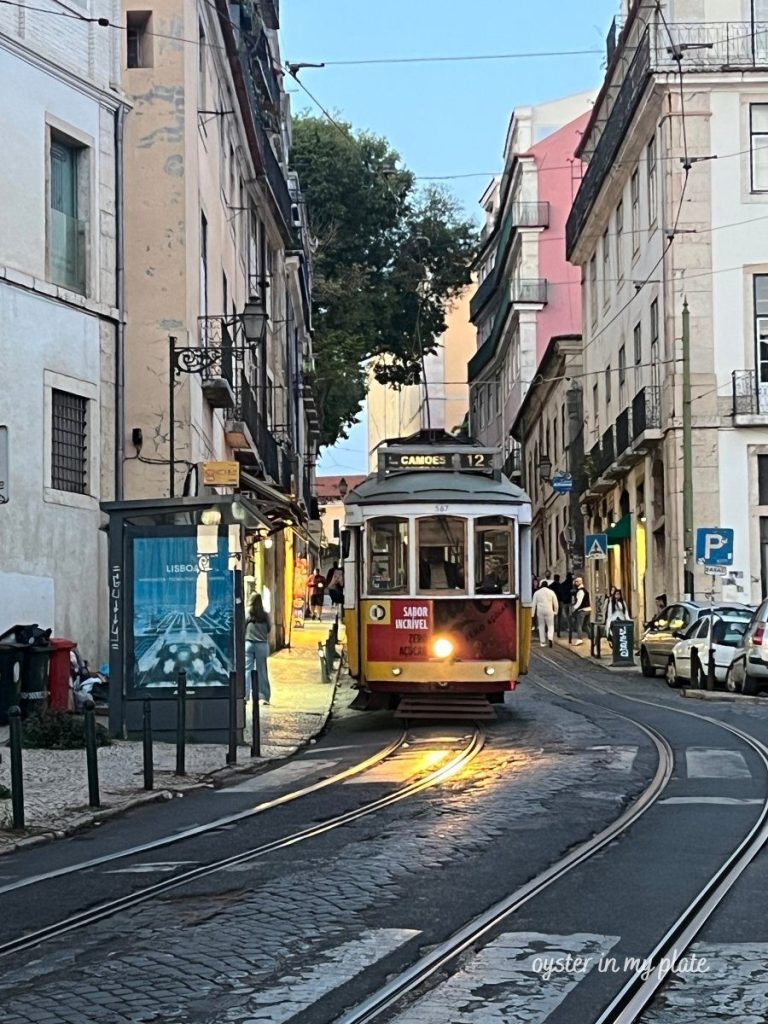 The historic bright yellow and red Tram 28 in Lisbon, seen on a narrow cobblestone street at dusk, with buildings and a tram stop nearby.