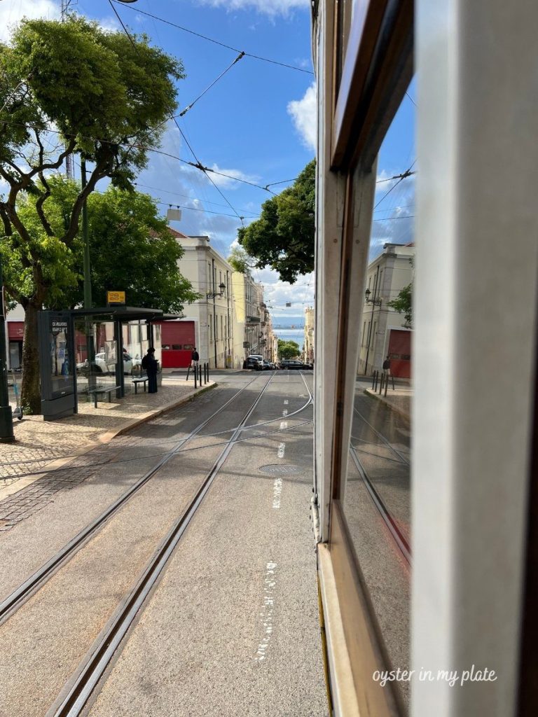 The historic bright yellow and red Tram 28 in Lisbon, seen on a narrow cobblestone street at dusk, with buildings and a tram stop nearby.