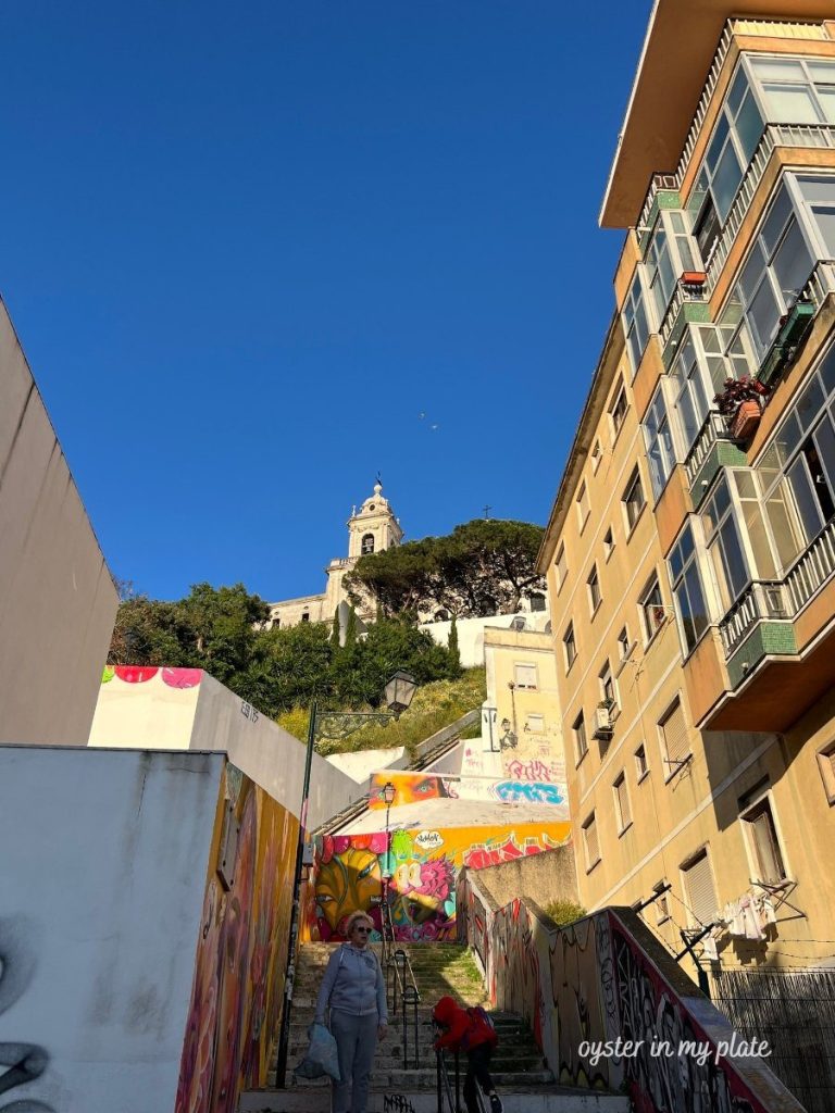 A wide outdoor staircase in Lisbon, adorned with colorful graffiti art, leading up towards a white church on a hill under a clear blue sky. A person walks up the stairs in the foreground.