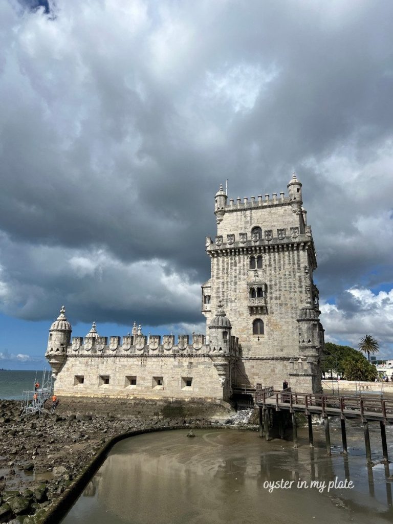 Belem Tower, a historic Portuguese fortress, under a dramatic cloudy sky with water and a wooden bridge in the foreground.