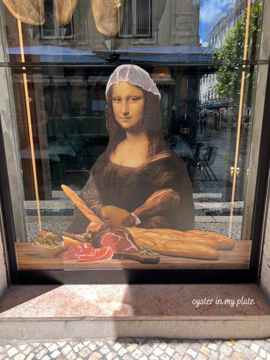A window display featuring a Mona Lisa parody wearing a hairnet, surrounded by baguettes, cured ham, and olives, in an outdoor setting