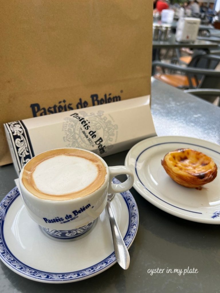 Pastéis de Belém coffee cup and saucer with an egg tart on a plate, and a paper bag in the background on an outdoor cafe table