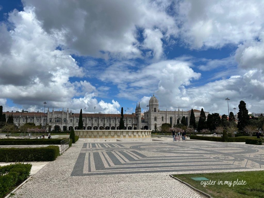 Jerónimos Monastery in Lisbon, a large stone monastery building with a patterned cobblestone plaza and trees under a cloudy sky